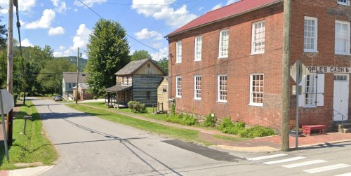 A quiet street scene featuring a brick building, a wooden house, and trees under a blue sky with fluffy clouds.