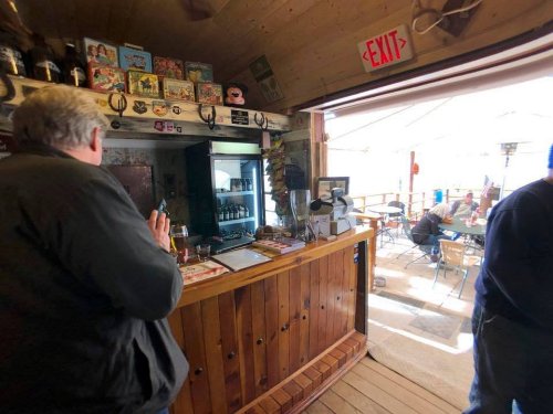 A rustic bar interior with a wooden counter, a man standing by the bar, and patrons seated outside under a canopy.