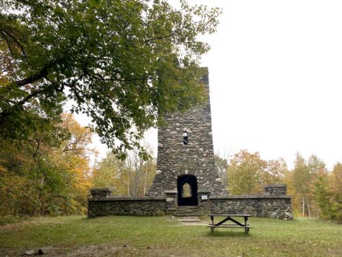 Stone tower surrounded by trees, with a picnic table in front and autumn foliage in the background.
