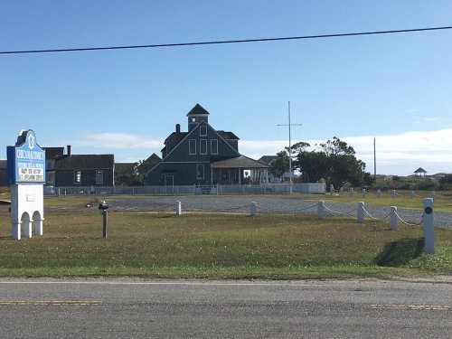 A large gray house with a tower and cross, surrounded by a grassy area and a white fence, under a clear blue sky.
