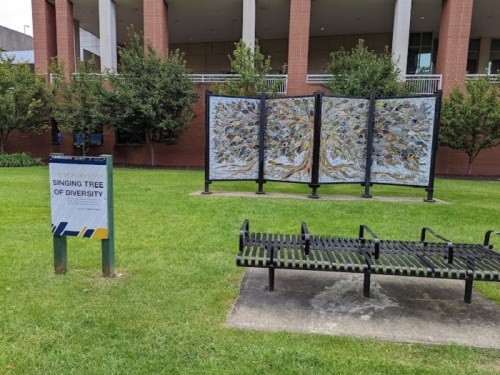 A park scene featuring a sign that reads "Singing Tree of Diversity" next to a colorful art installation and a bench.