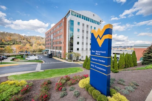 A modern building with a sign for One Waterfront Place and WVU Foundation, surrounded by landscaped greenery and blue skies.