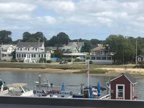 A scenic view of a marina with boats, sandy shore, and charming houses along the water under a partly cloudy sky.