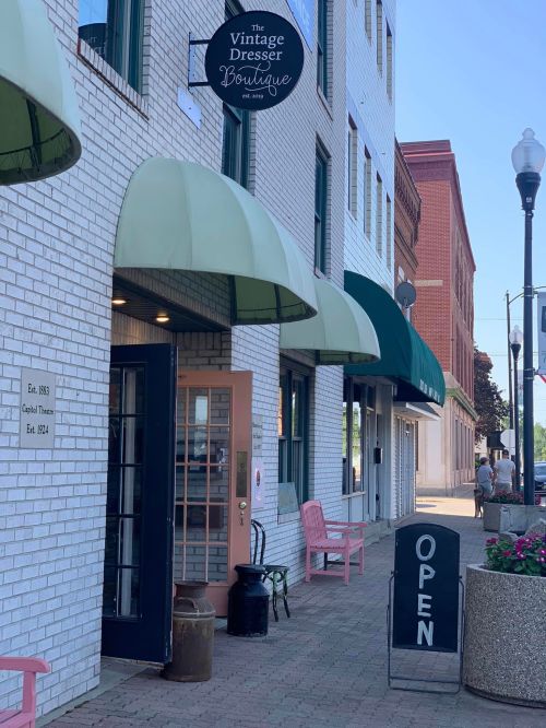 A boutique storefront with a green awning, open sign, and pink chairs outside, located on a city street.