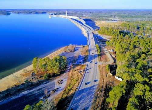 Aerial view of a serene lake with a road alongside, surrounded by trees and a clear blue sky.