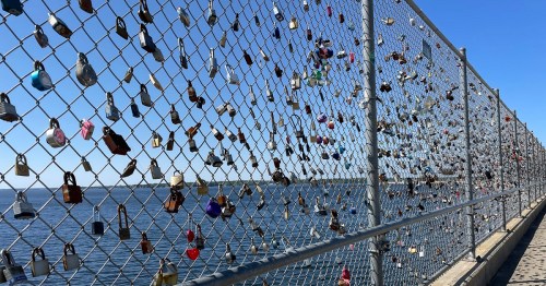A chain-link fence adorned with numerous colorful padlocks, overlooking a calm body of water under a clear blue sky.