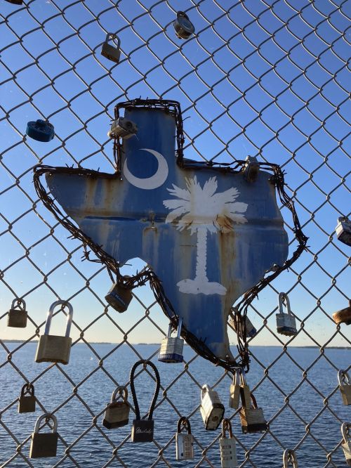 A Texas-shaped sign with a palm tree and crescent moon, surrounded by padlocks on a fence by the water.