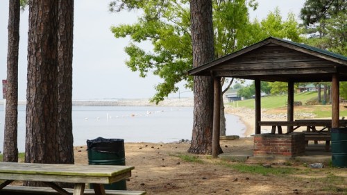A peaceful lakeside scene with a picnic shelter, trees, and a sandy beach in the background.