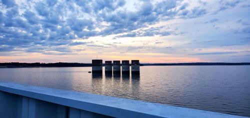 Sunset over a calm lake with a structure in the water and clouds in the sky, reflecting soft colors on the surface.