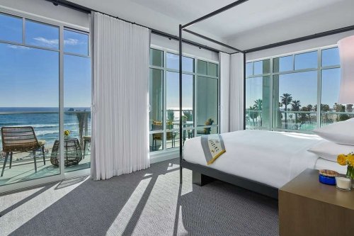 Bright hotel room with a four-poster bed, large windows, and ocean views, featuring modern decor and natural light.