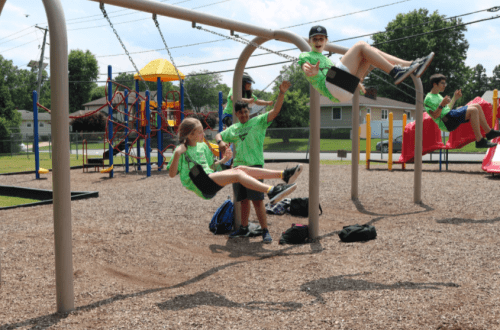 Children in green shirts swing on a playground, with colorful play structures in the background on a sunny day.
