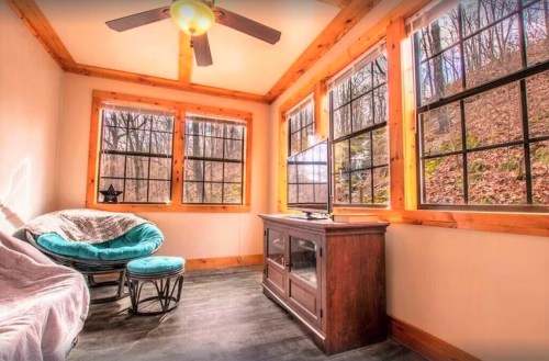 Cozy sunlit room with large windows, a ceiling fan, a TV, and a comfortable chair. Surrounded by trees outside.