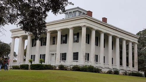 A historic white mansion with tall columns, surrounded by green grass and trees, under a cloudy sky.