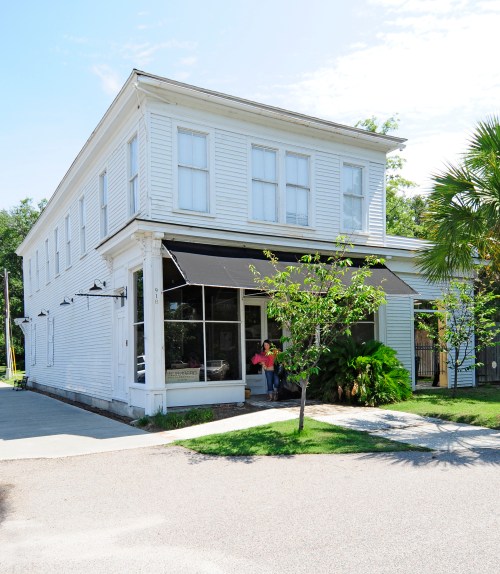 A white two-story building with large windows and an awning, surrounded by greenery and a sidewalk.