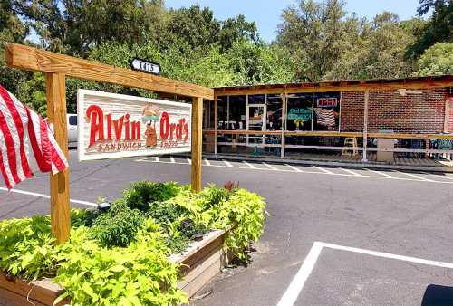 A sign for Alvin & Orie's Sandwich Shop in front of a brick building with outdoor seating and greenery.