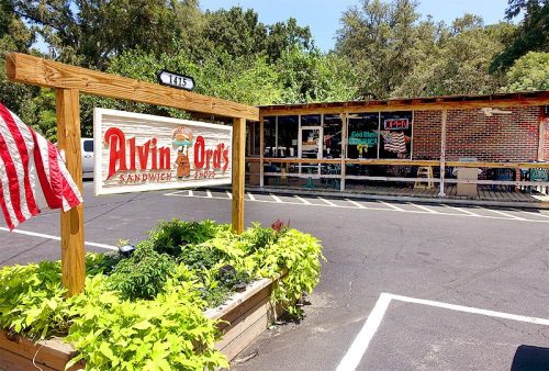 A sign for Alvin & Orie's Sandwich Shop in front of a brick building with outdoor seating and greenery.
