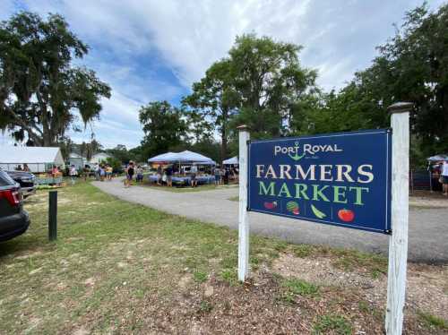 A sign for Port Royal Farmers Market with tents and people browsing in a green park setting.