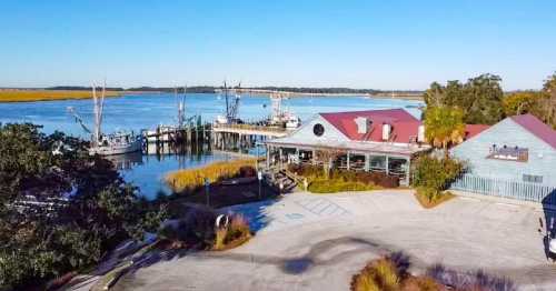 A scenic view of a marina with fishing boats, a restaurant, and calm waters under a clear blue sky.