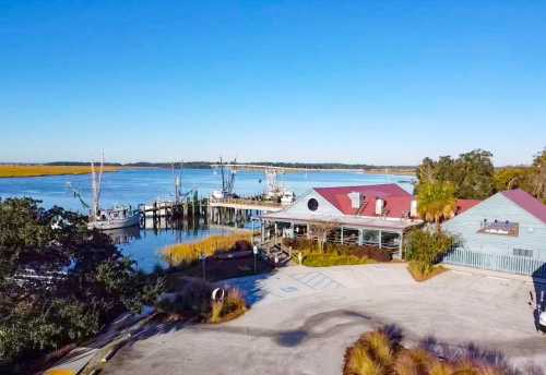 A waterfront restaurant with boats docked nearby, surrounded by marshland and clear blue skies.