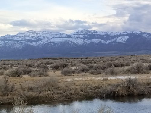 Snow-capped mountains rise above a grassy landscape, with a calm river reflecting the sky in the foreground.