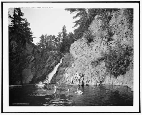 Black and white photo of people swimming in a natural pool near a waterfall, surrounded by rocky cliffs and trees.