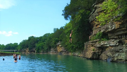 A person swings from a rope off a cliff into a river, while others swim nearby on a sunny day.