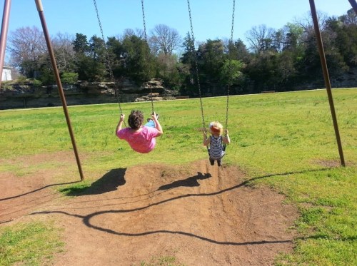 Two children swing on swings in a grassy park, surrounded by trees and a rocky backdrop.