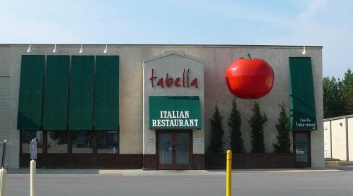 Exterior of Tabella Italian Restaurant featuring a large red tomato sign and green awnings.