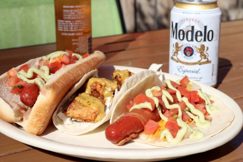 A plate with two hot dogs topped with sauce, tacos with bacon and toppings, and a can of Modelo beer.
