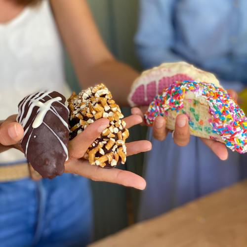 A person holds three colorful dessert tacos topped with sprinkles, pretzels, and chocolate drizzle.