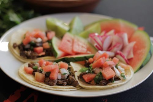 A plate of three tacos filled with beef, topped with diced tomatoes and onions, alongside slices of watermelon and cucumber.