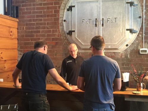 Two men stand at a wooden counter, engaging with a staff member in a casual, rustic setting.