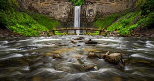 A scenic waterfall cascades down rocky cliffs, surrounded by lush greenery, with a bridge and people in the foreground.