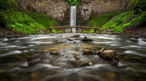 A scenic waterfall cascades down rocky cliffs, surrounded by lush greenery and a flowing river with smooth stones.