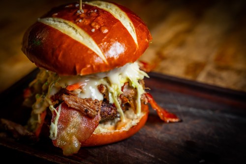 A close-up of a pretzel bun burger filled with pulled meat, bacon, and coleslaw, served on a wooden platter.