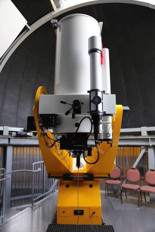 A large telescope inside an observatory, featuring a yellow base and a cylindrical white body, with chairs in the background.