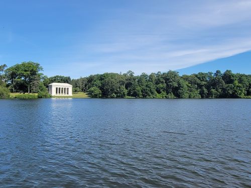 A serene lake scene with a classical white structure and lush green trees along the shore under a clear blue sky.