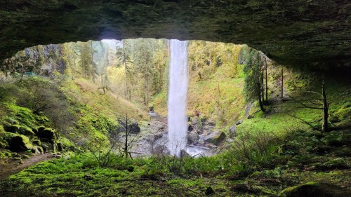 A waterfall cascades down into a lush green valley, framed by a rocky overhang and surrounded by trees.