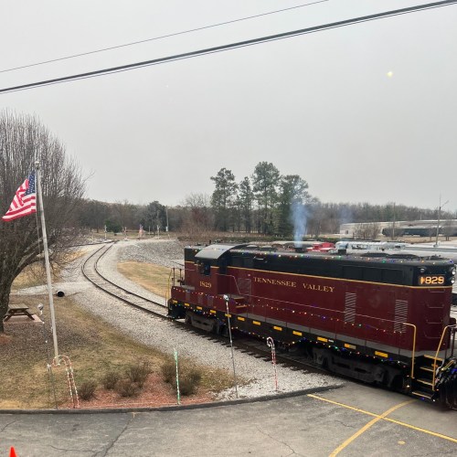 A maroon train engine sits near a flag, with tracks curving into the distance on a cloudy day.