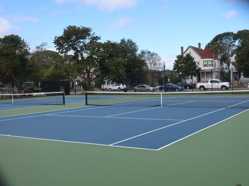 A view of empty tennis courts with a house in the background and trees lining the area under a blue sky.