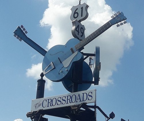 Sign featuring a blue guitar and a highway marker for Routes 61 and 49, labeled "The Crossroads," against a cloudy sky.