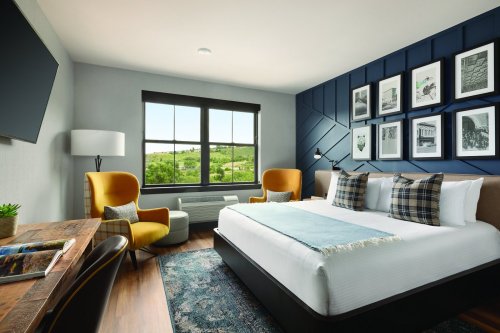 A modern hotel room featuring a queen bed, two armchairs, a desk, and large windows with scenic views.