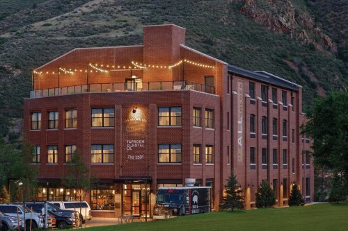 A brick hotel with string lights on the rooftop, surrounded by green hills and trees, in a scenic outdoor setting.