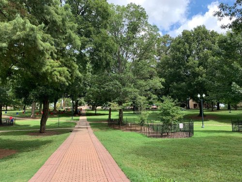 A paved path winds through a lush green park with trees and a fenced area, under a partly cloudy sky.
