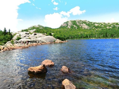 A serene lake surrounded by rocky shores and lush green hills under a bright blue sky with scattered clouds.