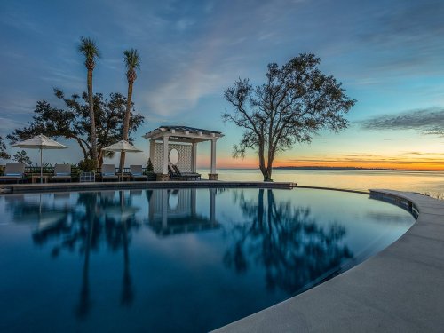 A serene poolside scene at sunset, featuring palm trees, lounge chairs, and a gazebo reflecting in the water.