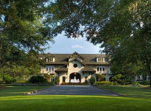 A large, elegant house surrounded by lush greenery and trees, with a paved pathway leading to the entrance.