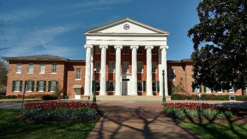 Historic building with white columns, red brick facade, and landscaped flower beds under a clear blue sky.