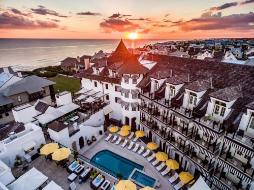 Aerial view of a coastal resort at sunset, featuring a pool, lounge chairs, and a scenic ocean backdrop.