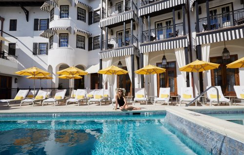 A woman in a black swimsuit sits by a pool surrounded by yellow umbrellas and lounge chairs at a stylish hotel.